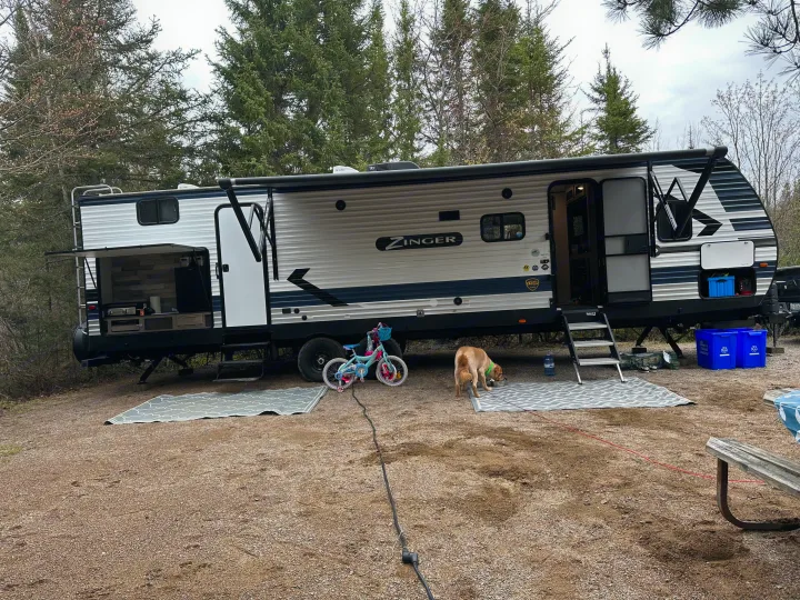 A Zinger RV rental is set up at a scenic campsite with an outdoor kitchen, a playful dog, and a child's bike.. Crossroads RV Zinger 2021