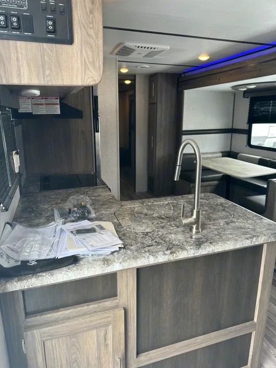 Inside an RV rental: a bright kitchen with a modern sink, stovetop, and overhead control panel. Dinette and hallway are visible, with manuals on the grey counter.. Crossroads RV Zinger 2021
