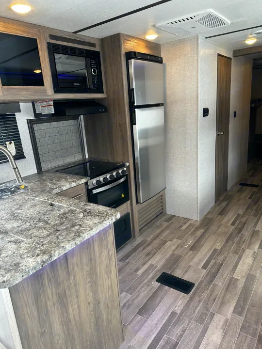 Modern RV rental kitchen featuring a microwave, refrigerator, stove/oven, sink, and gray wood-look flooring for comfort on the road.. Crossroads RV Zinger 2021