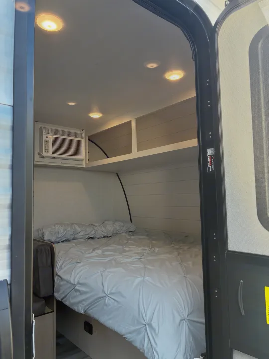 Light-colored RV rental interior featuring a bed with gray ruched bedding, AC unit, and overhead storage, visible from the open door.. Jayco 1UJBC0AJ6F7750334 2026
