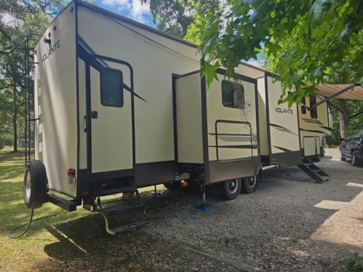 Cream Volante travel trailer rv rental with extended slides and awning at a gravel campsite under trees.. Crossroads RV Volante 2020