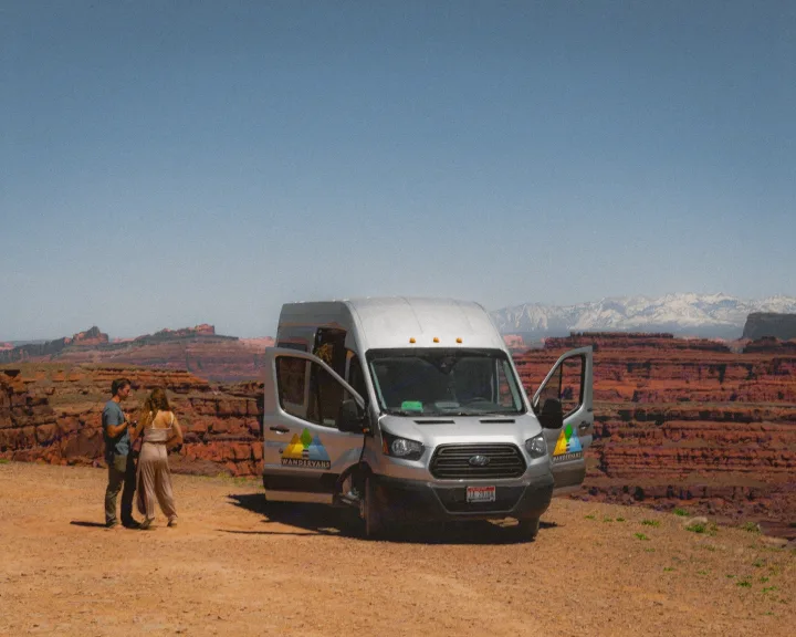 A couple stands by their silver RV rental van in a vast desert landscape, with red rock formations and snowy mountains.. Ford Transit Custom 2022