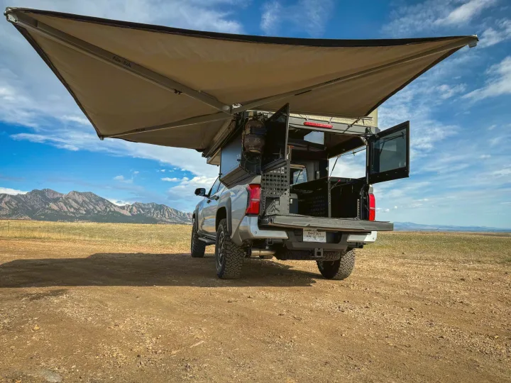 Overland truck camper, an ideal RV rental for off-grid camping adventures. Awning extended in a scenic field with mountains.. Toyota Tacoma 2026