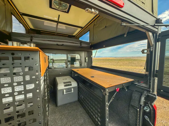 Compact RV rental camping setup in a truck bed with a pop-top camper, portable fridge, and fold-out table, overlooking an open landscape.. Toyota Tacoma 2026