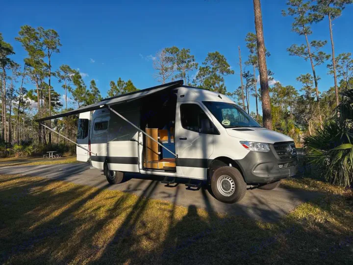RV rental adventure: a white Mercedes Sprinter camper van with an awning extended at a sunny campsite among tall trees.. Mercedes-Benz Sprinter 2023