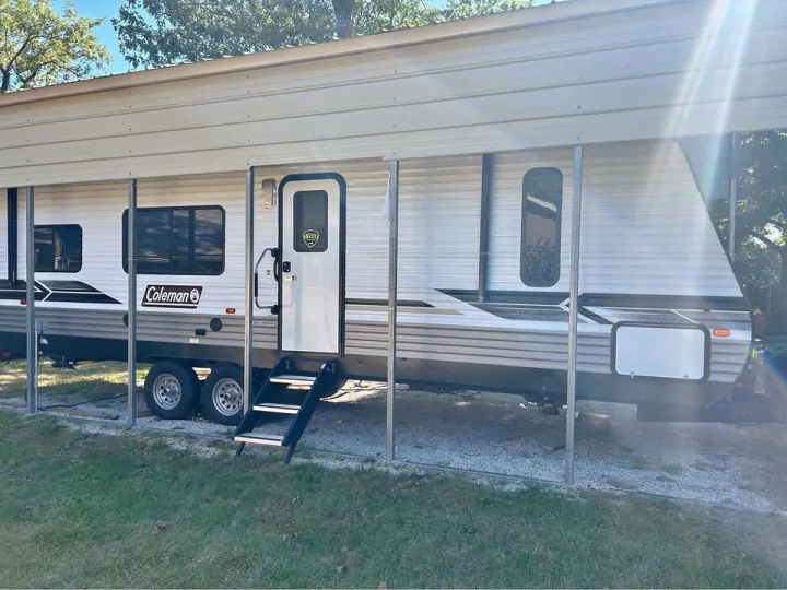 Coleman travel trailer RV rental parked under a metal carport. Entry steps are extended over gravel.. Coleman 270TQ 2022