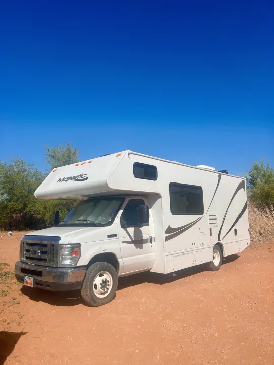 A white Majestic Class C RV rental is parked on reddish dirt under a clear blue sky, with sparse green bushes in the background.. Thor Majestic 2018