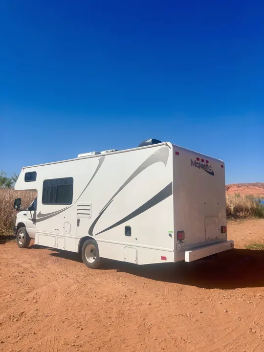 A white Majestic **rv rental** is parked on reddish desert ground under a clear blue sky, ideal for your next outdoor adventure.. Thor Majestic 2018