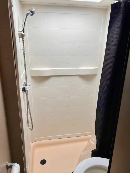RV rental shower: a compact unit with white tiled walls, handheld showerhead, shelf, peach pan, and a black shower curtain.. Thor Majestic 2018