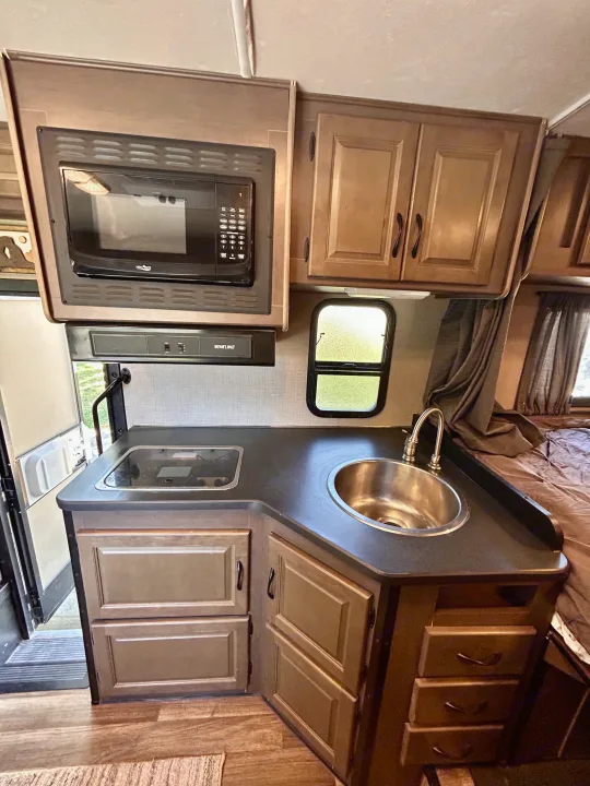 Inside an RV rental: a compact kitchen with microwave, stovetop, sink, and wood cabinets, ready for your next adventure.. Thor Majestic 2018