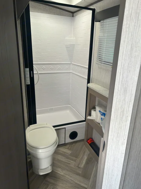 Compact RV rental bathroom features a toilet, white subway-tile-look shower, and shelving with essentials.. Cruiser RV Shadow Cruiser 2022