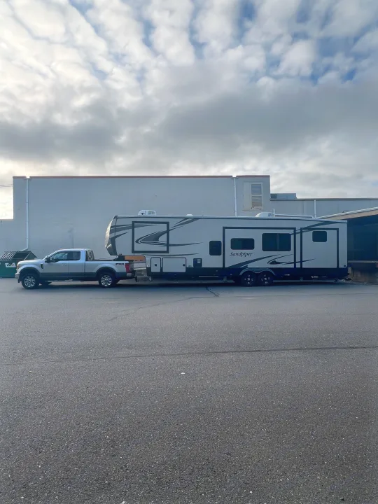 Silver pickup truck towing a large Sandpiper fifth-wheel RV for an rv rental. Parked near a building under cloudy skies.. Forest River Sandpiper 2021
