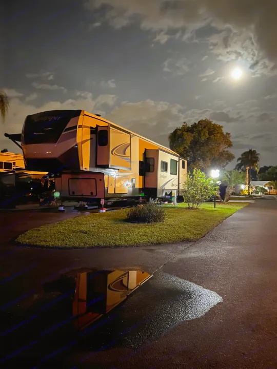 A cozy RV rental, illuminated under a cloudy moonlit sky, reflects in a roadside puddle at a serene campground.. Forest River Sandpiper 2021