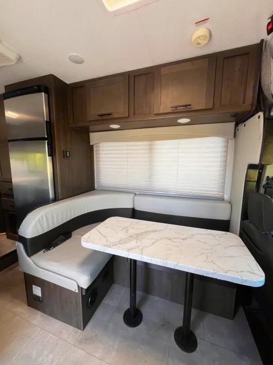 RV rental interior: a modern U-shaped dinette with gray seating, a white marble-look table, and an adjacent refrigerator.. Coachmen Cross Trail 2023