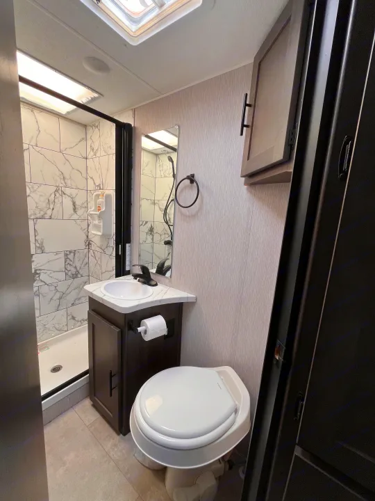 RV rental bathroom showing a white tiled shower, dark wood vanity with sink, white toilet, and textured grey walls.. Coachmen Cross Trail 2023