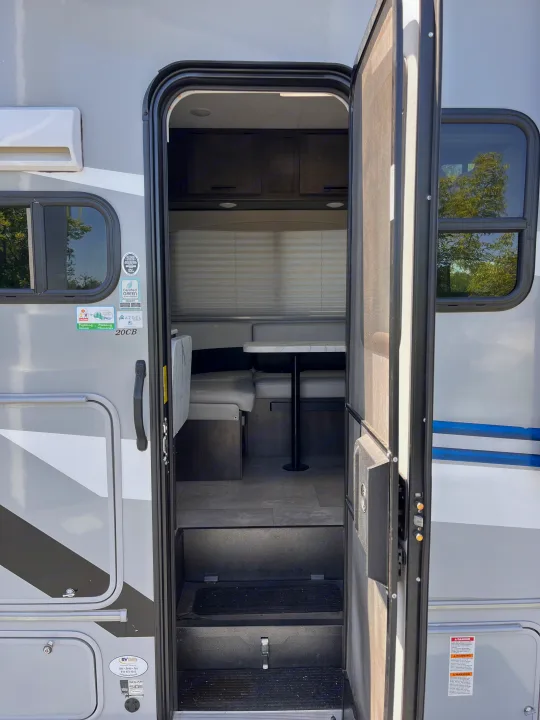 Open door of an **rv rental** showing a compact dinette, interior steps, and exterior decals including the "20CB" model.. Coachmen Cross Trail 2023