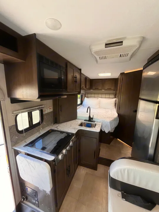 RV rental interior with a modern kitchen: marble-look counter, stove, sink, and microwave. A comfy bed is visible in the back.. Coachmen Cross Trail 2023
