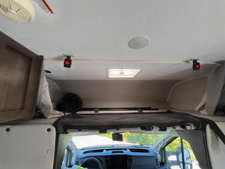 Overhead bunk bed in an RV rental, featuring a safety net and ceiling fixtures above the vehicle's dashboard.. Coachmen Cross Trail 2023