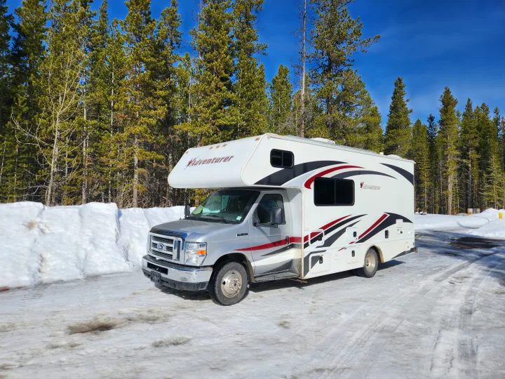 Silver and white Adventurer RV rental on snowy ground with tall pine trees under a clear blue sky, next to large snowbanks.. Adventurer Adventurer Motorhome 2017