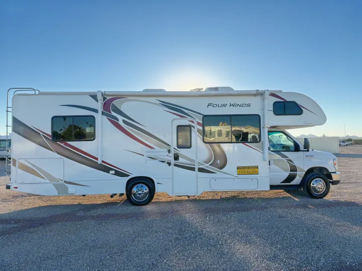 A white Four Winds RV rental with dark graphics is parked on a gravel lot under a clear blue sky, ideal for travel.. Thor Four Winds 2019