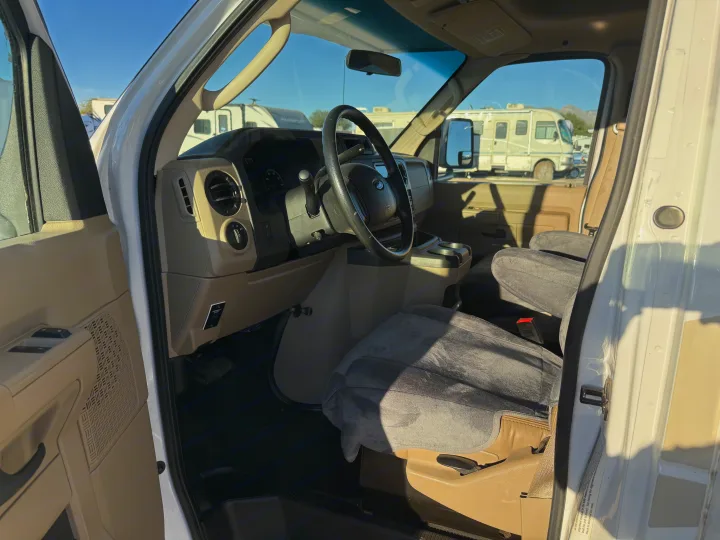 RV rental van's driver-side interior with steering wheel, dashboard, and soft-covered seats. Other RVs are visible outside.. Thor Four Winds 2019