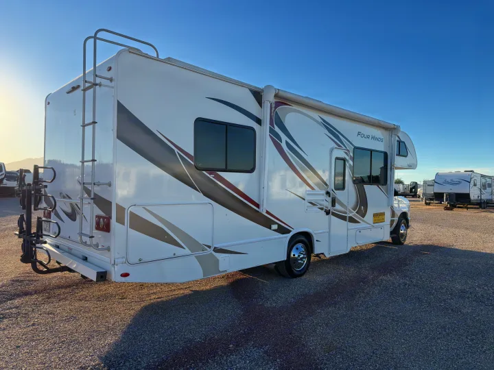 White Four Winds RV rental with striking graphics, a ladder, and bike rack, parked on a sunlit gravel lot.. Thor Four Winds 2019