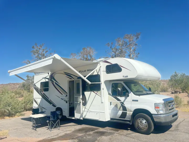 RV rental: A white Four Winds motorhome with its awning extended is parked at a sunny campsite with chairs and a table.. Thor Four Winds 2019
