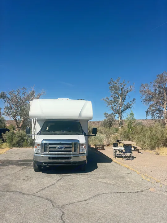 A white Ford RV rental is parked at a sunny campsite with outdoor furniture and a grill, surrounded by sparse trees.. Thor Four Winds 2019
