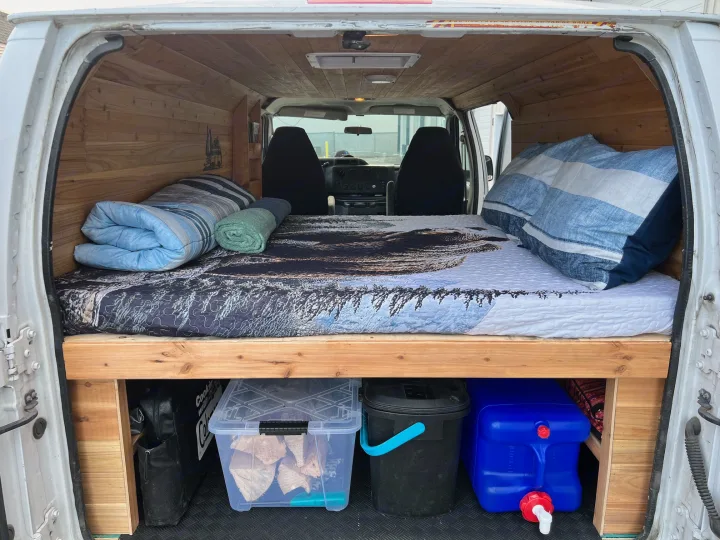 Inside an RV rental van: a cozy bed with pillows, wood paneling, and storage bins under the bed platform, ready for travel.. Ford Econolline 250s 2013