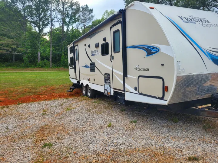 Tan Coachmen Freedom Express travel trailer (RV rental) parked on gravel next to a grassy area with trees.. Coachmen Freedom Express 2019