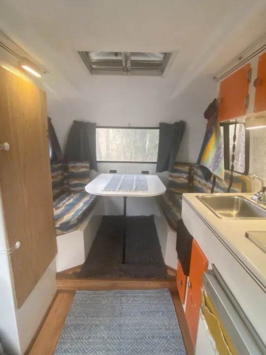 This cozy RV rental interior features a dinette with patterned cushions, a compact kitchen, and a window revealing trees.. Scamp 13' 1987