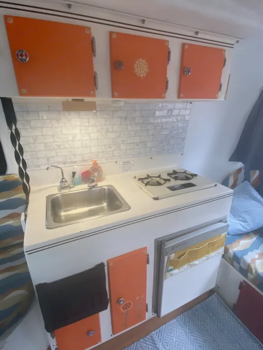 Cozy RV rental kitchen with vibrant orange cabinets, subway tile backsplash, sink, stove, and compact fridge. Perfect for road trips.. Scamp 13' 1987