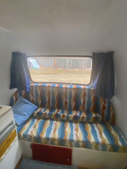 Cozy rv rental interior featuring a patterned bench/bed, window view of trees, and blue pillow. Ideal for comfortable travel.. Scamp 13' 1987