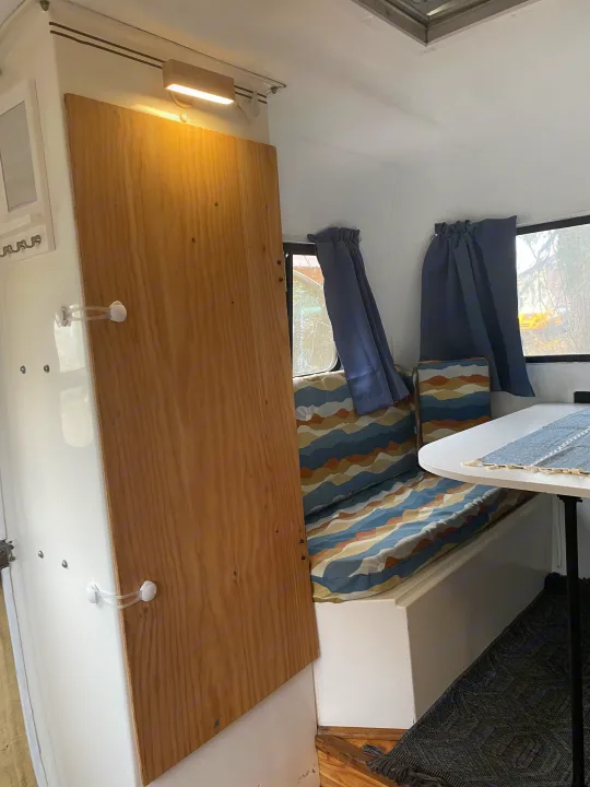 Interior of an rv rental: a light wood partition, patterned bench seating, dining table, and curtained windows create a cozy space.. Scamp 13' 1987