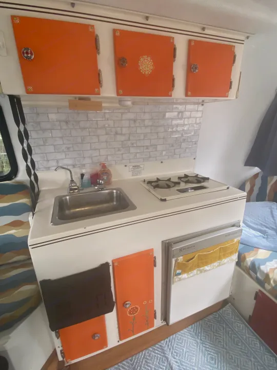 Bright orange cabinets and white appliances in a retro-themed RV rental kitchen, with a sink, stove, and tile backsplash.. Scamp 13' 1987
