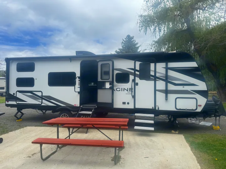 This rv rental, a white and black Grand Design travel trailer, is set up at a campsite with a red picnic table.. Grand Design Imagine 2024