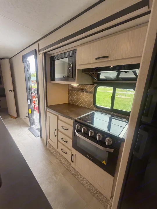 Functional rv rental kitchen featuring a black microwave, stove/oven, dark counters, and light wood cabinets. Ideal for camping.. Coachmen Apex 2021