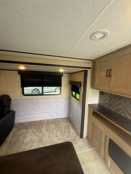 This RV rental shows a clean interior with wood-look cabinets, a gray herringbone backsplash, carpet, and windows.. Coachmen Apex 2021