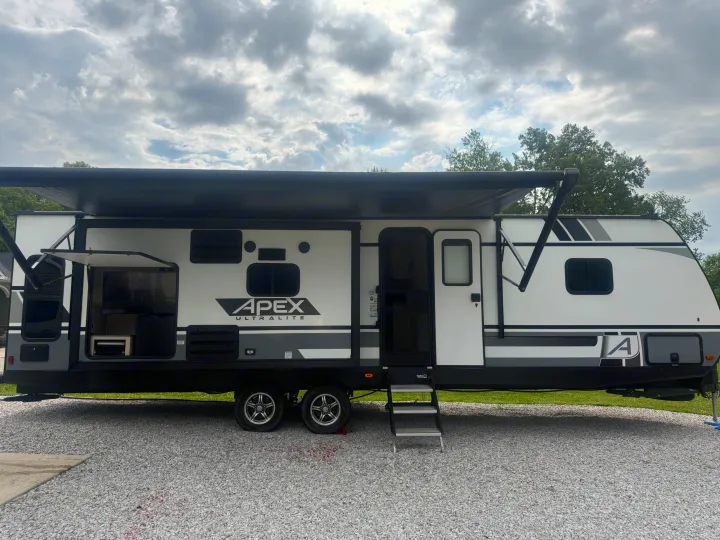 A modern white and gray Apex Ultralite RV rental with an extended awning, open outdoor kitchen, and entry steps on gravel.. Coachmen Apex 2021