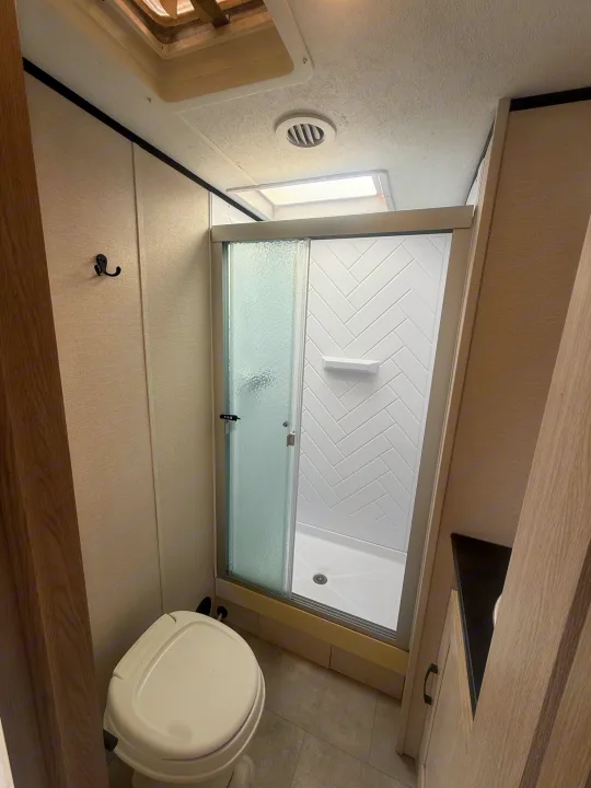 Compact RV rental bathroom featuring a toilet, sliding glass shower with herringbone tile, and a wall hook.. Coachmen Apex 2021