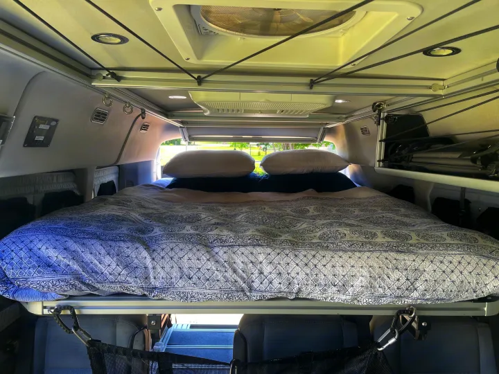 Inside an RV rental: a cozy bed with patterned bedding, overhead storage system, and fan for comfortable van life travel.. Ford Transit 2018