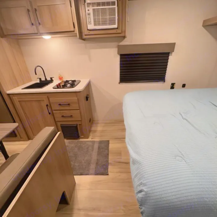 A bright interior view of an RV rental showing a compact kitchen with a sink and stove, an air conditioning unit, and a bed with a light blue cover.. Keystone RV Coleman 2025