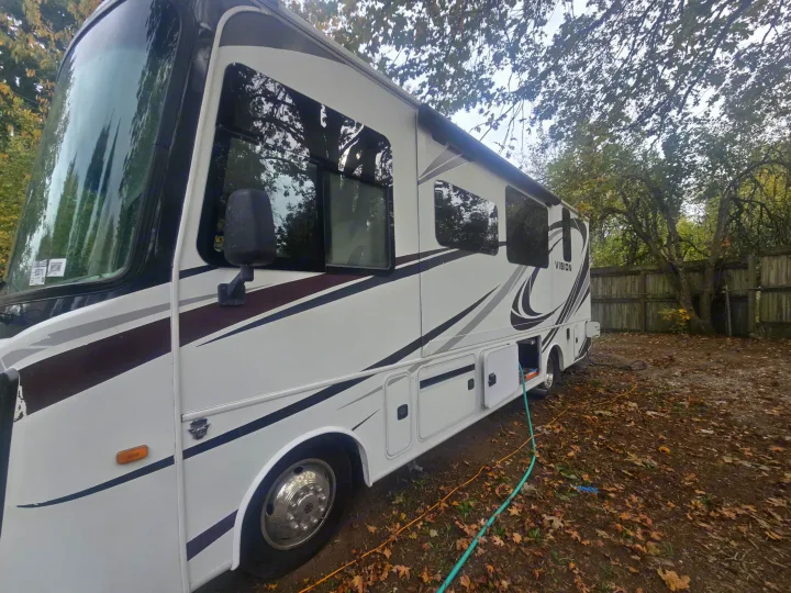 This Class A RV rental, a white motorhome with a deployed awning, is parked outside with hookups on a leafy ground.. Entegra Coach Vision 2019