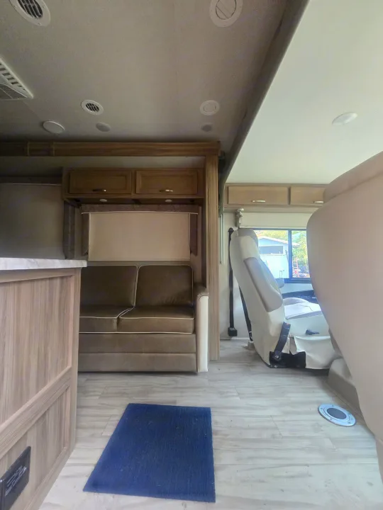 Inside an RV rental: a brown sofa, wood cabinets, beige captain's chair, light wood-look floor, and ceiling vents.. Entegra Coach Vision 2019