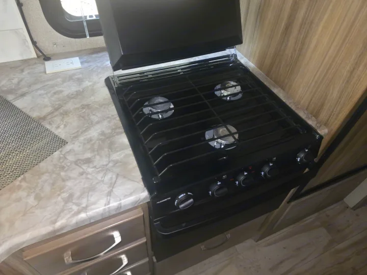 This RV rental kitchen features a black Dometic gas stovetop and oven, integrated into a light marbled countertop, complete with controls and a lid.. Entegra Coach Vision 2019