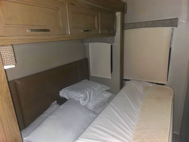 RV rental bedroom with a bed, white linens, padded headboard, wood overhead cabinets, and light-colored window blinds.. Entegra Coach Vision 2019