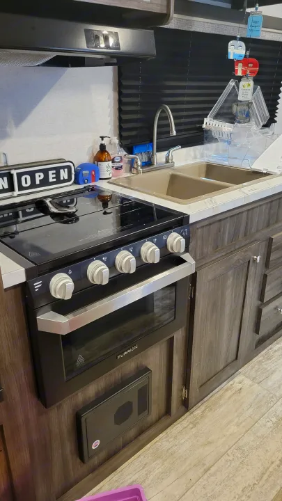 RV rental kitchen: black Furrion stove/oven, double sink, soap bottles, wood-grain cabinets, and a 'Happy Campers' mobile.. Highland Ridge RV Open Range 2020