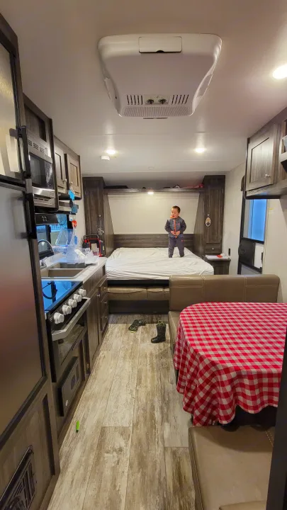 An RV rental interior showing a child on the bed, a kitchen with a stove/sink, and a dining area with a red checkered table.. Highland Ridge RV Open Range 2020