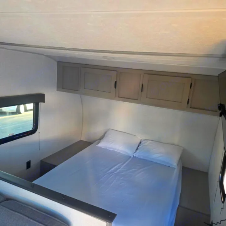 RV rental bedroom with a neat white bed, overhead cabinets, and a window, offering comfortable sleeping space in a compact setup.. Clipper 5K 2024