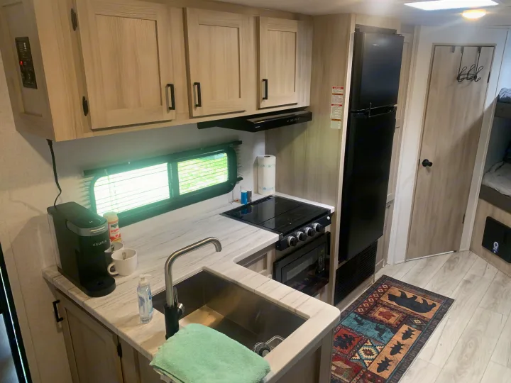 Compact rv rental kitchen with light wood cabinetry, sink, stove, microwave, fridge, Keurig, and colorful rug.. Forest River East to West 2025